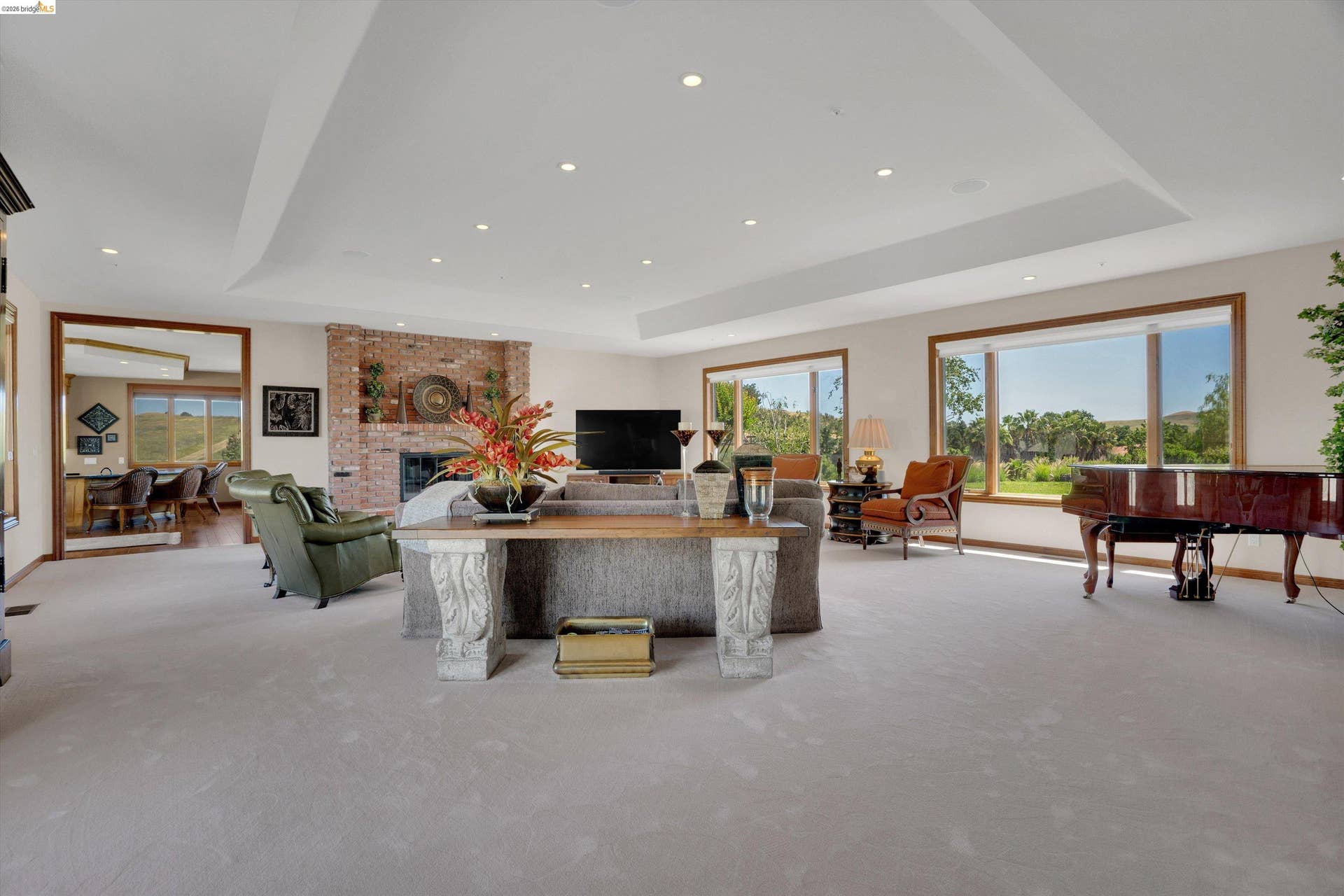 Spacious living area featuring expansive windows, a brick-faced fireplace, recessed lighting, a tray ceiling, and light-toned carpeting