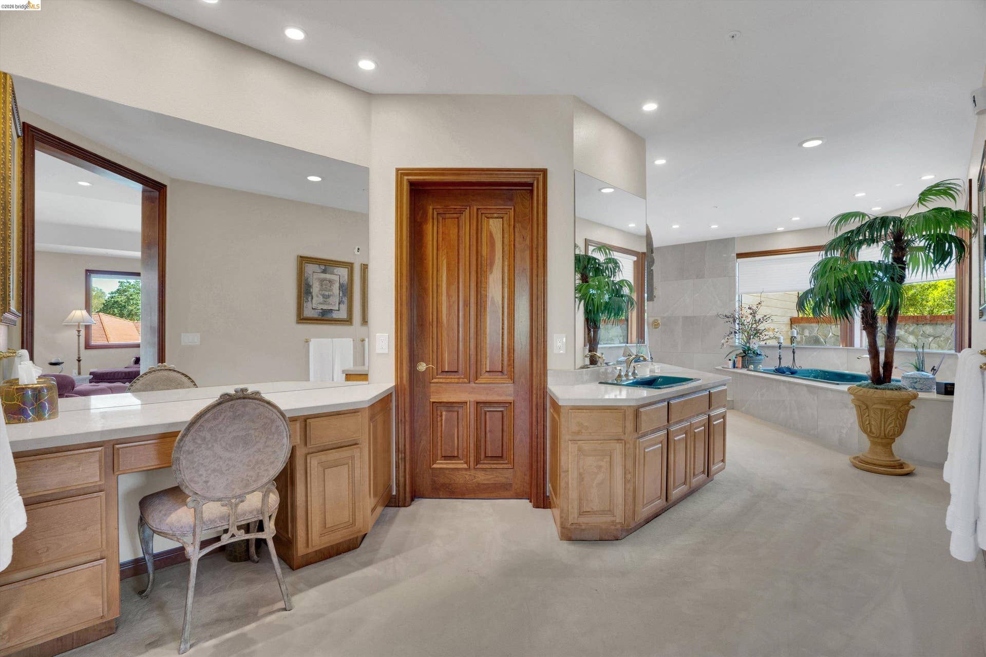 Spacious bathroom featuring a built-in vanity, light wood cabinetry, white countertops, dual sinks with chrome fixtures, and a large soaking tub