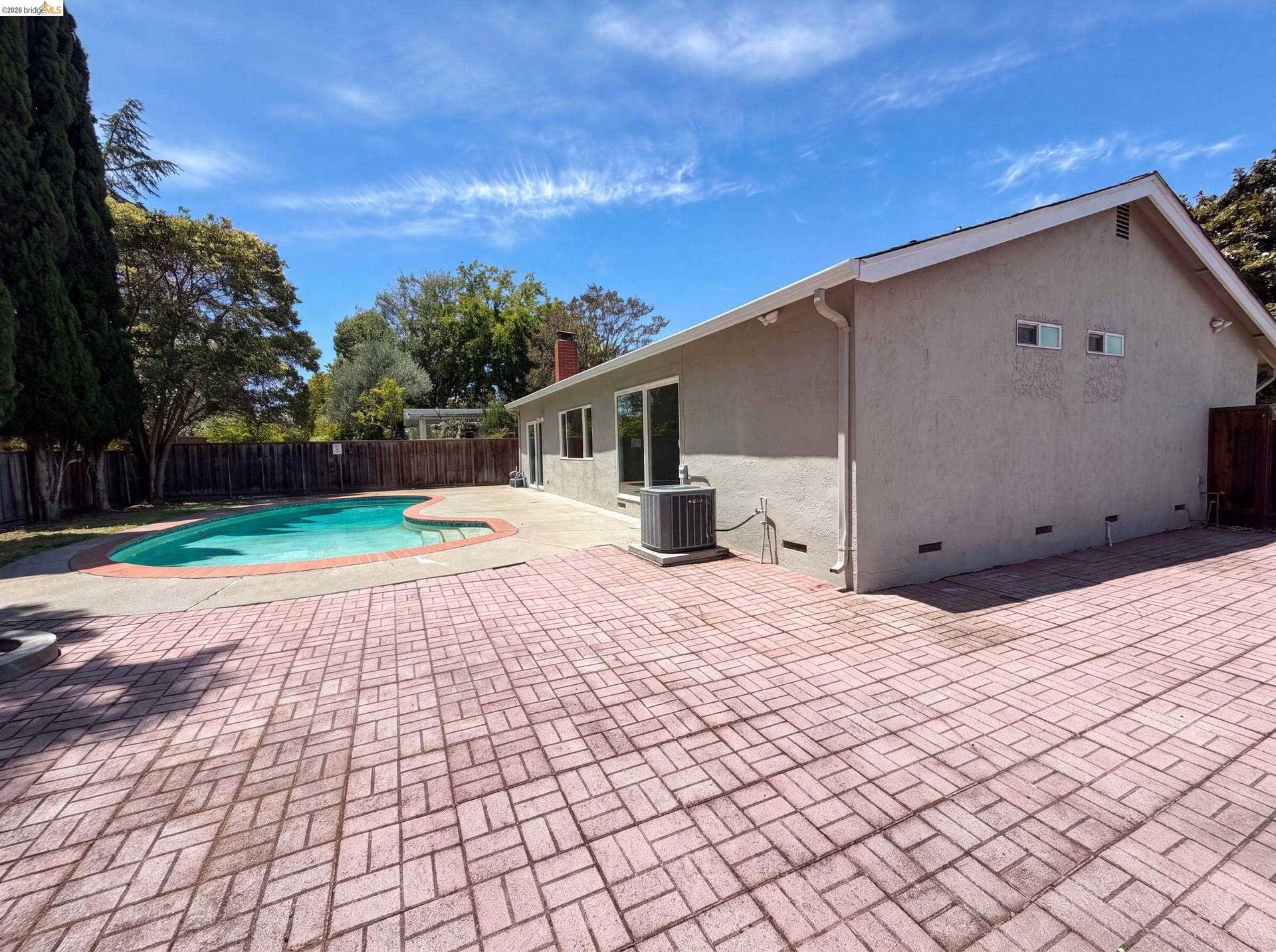 Paved patio featuring a kidney-shaped pool, mature perimeter trees, wood fencing, and a stucco exterior