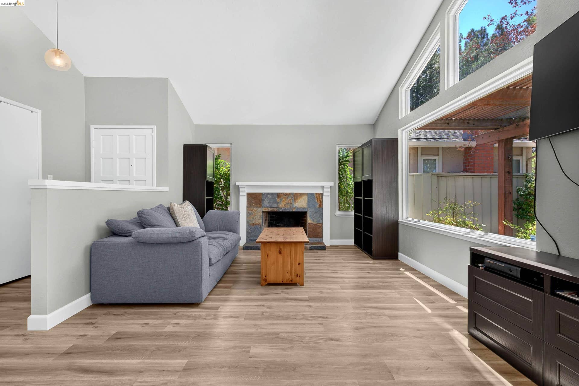 Living area featuring vaulted ceilings, wood-finish flooring, a stone-faced fireplace with a white mantel, and large picture windows