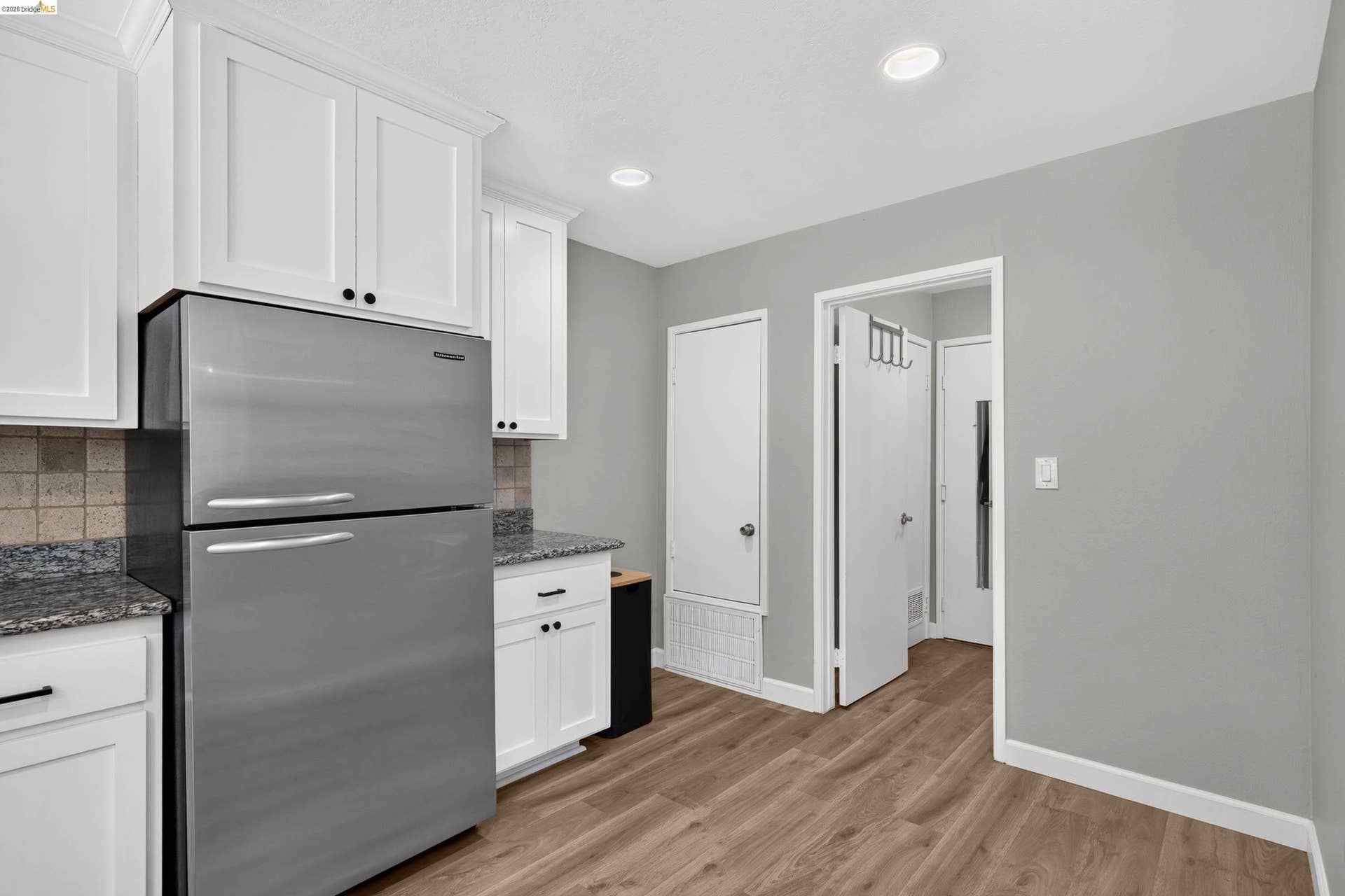 Kitchen featuring wood-finish flooring, white cabinetry with dark hardware, a stainless steel refrigerator, and granite countertops with a tiled backsplash