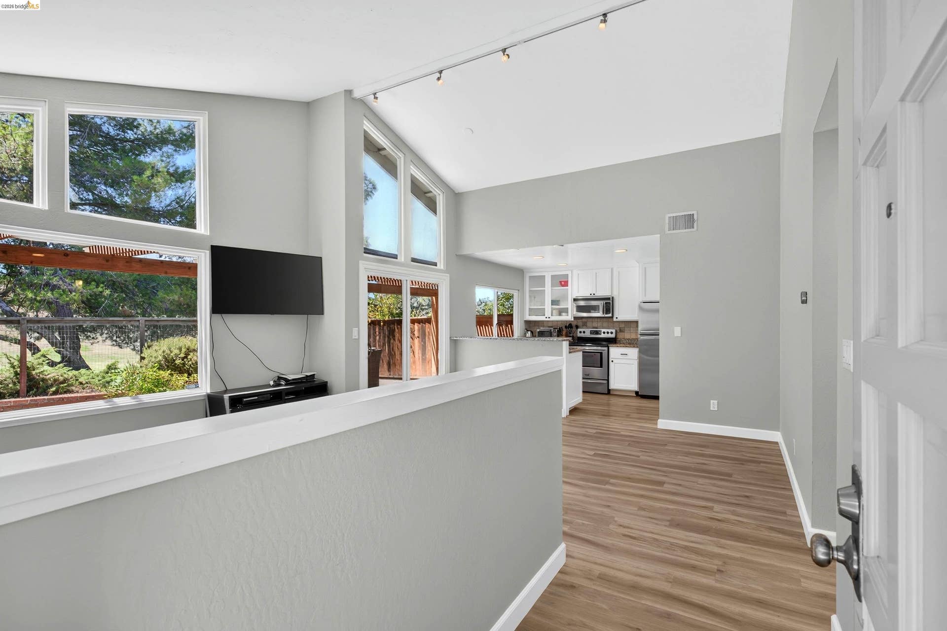 Vaulted ceiling with track lighting, wood-finish flooring, ample natural light from large windows and sliding glass doors, white kitchen cabinetry with stainless steel appliances
