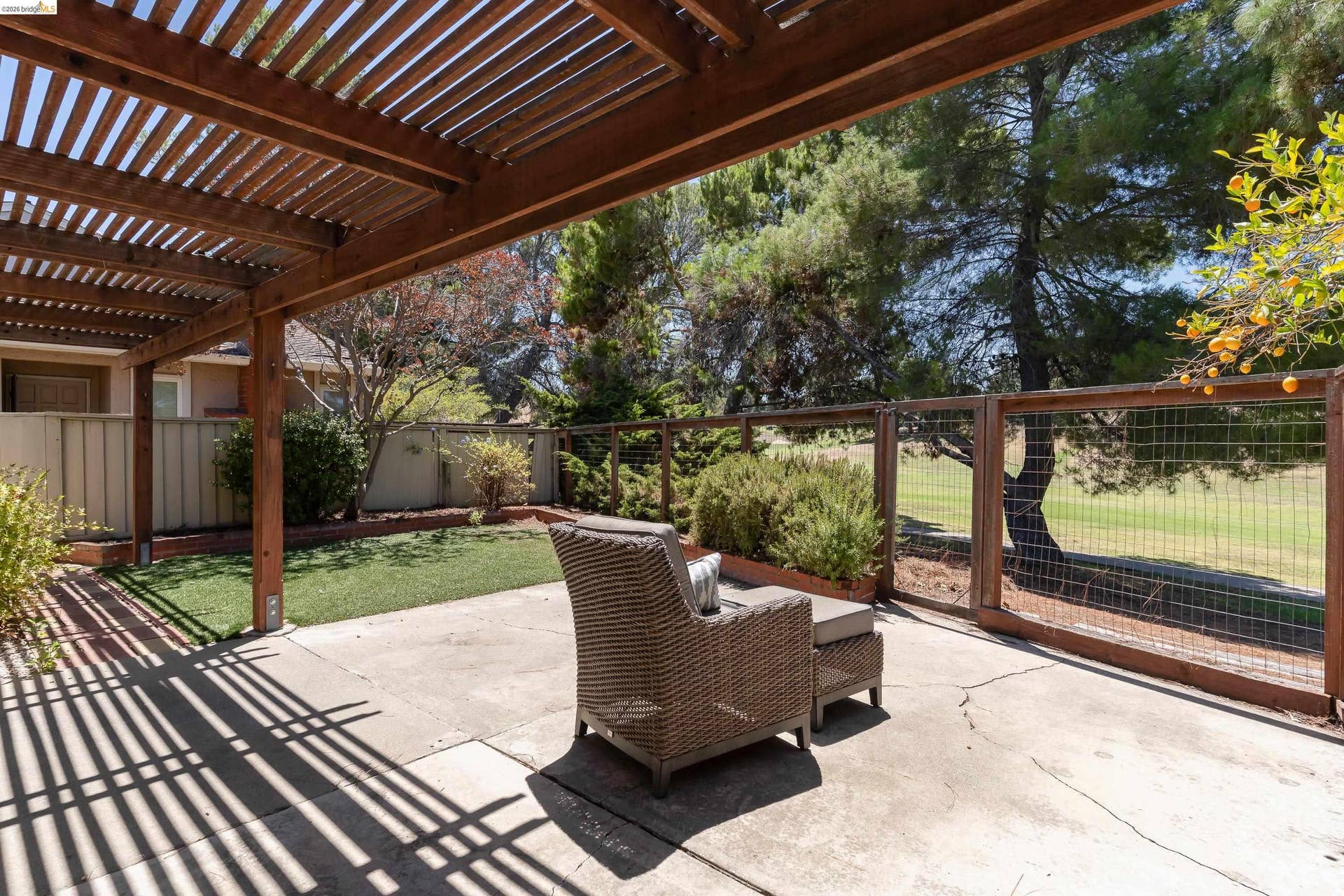 Patio area featuring a wood pergola providing partial shade, concrete flooring, and a fenced backyard with artificial turf and mature landscaping