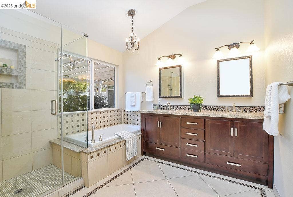 Glass-enclosed shower with tiled walls and mosaic-tiled niches, built-in soaking tub with mosaic-tiled surround, dual vanity with wood-finish cabinetry, light-toned countertops, and dual framed mirrors, and recessed lighting