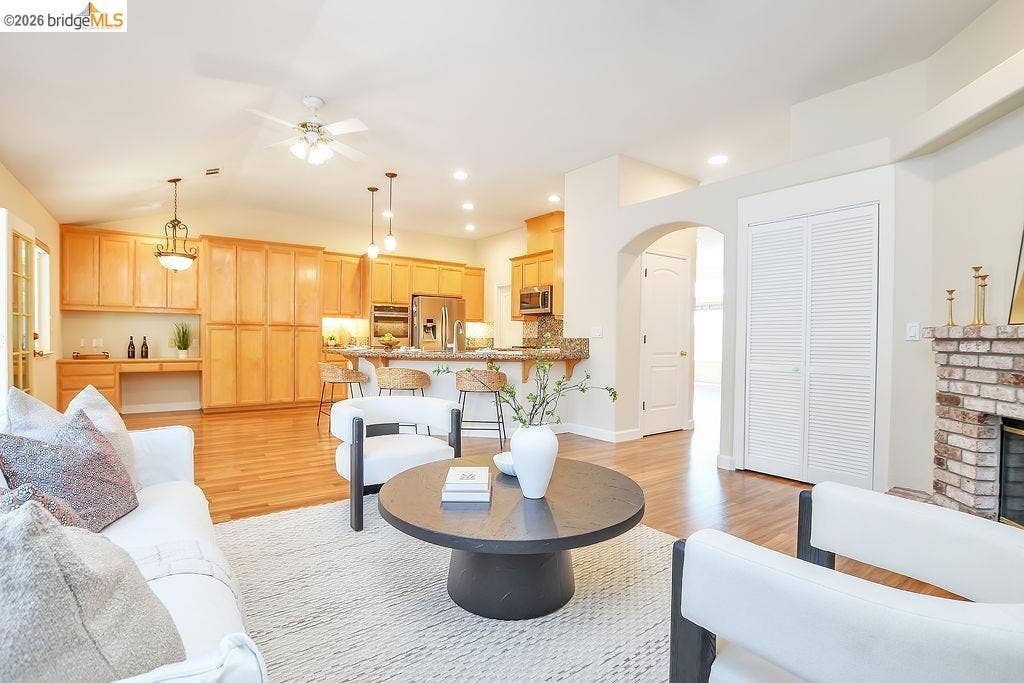 Vaulted ceilings, wood-finish flooring, kitchen with light wood cabinetry, stone countertop island, and a brick fireplace