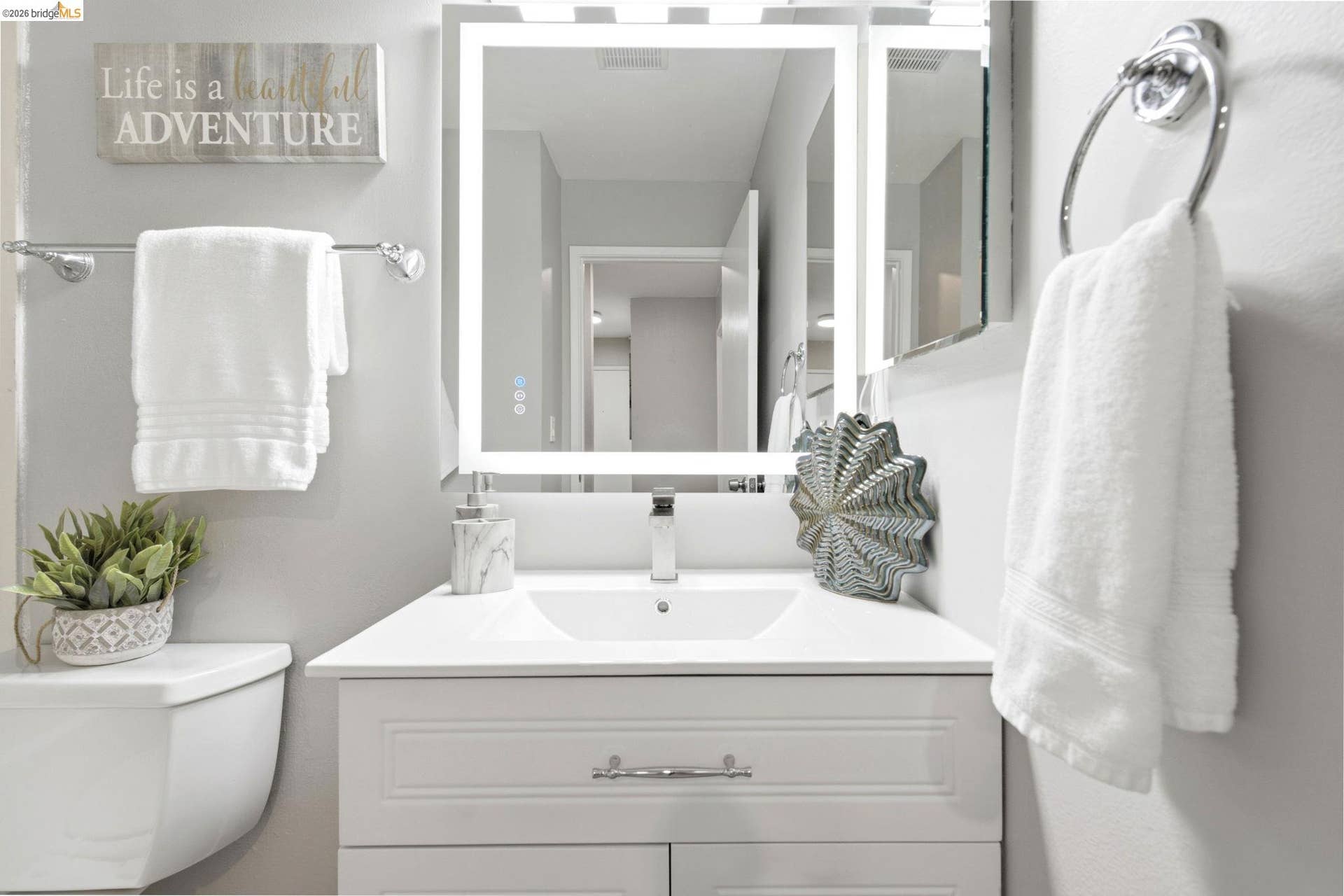 Bathroom vanity with a lighted mirror, polished chrome fixtures, white paneled cabinetry, and a white countertop with an integrated sink