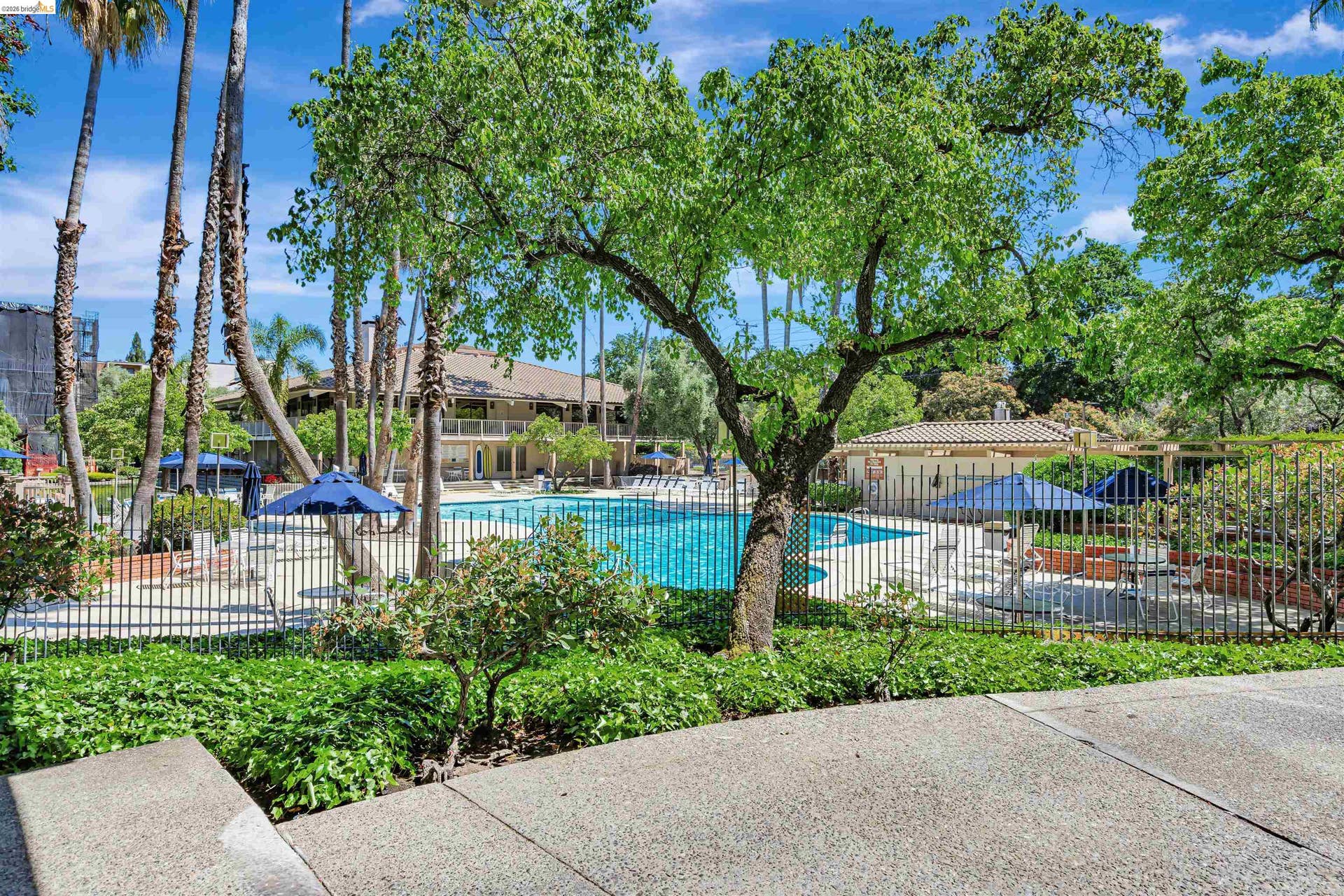 Spacious outdoor pool area featuring a perimeter fence, a pool house with a tile roof, mature trees, and surrounding landscaping